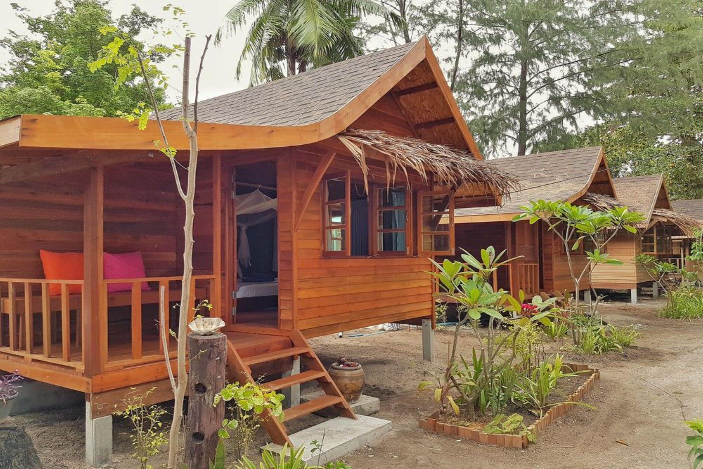 Una cabaña de madera con porche, techo de paja y un jardín con plantas y una pequeña fuente.
