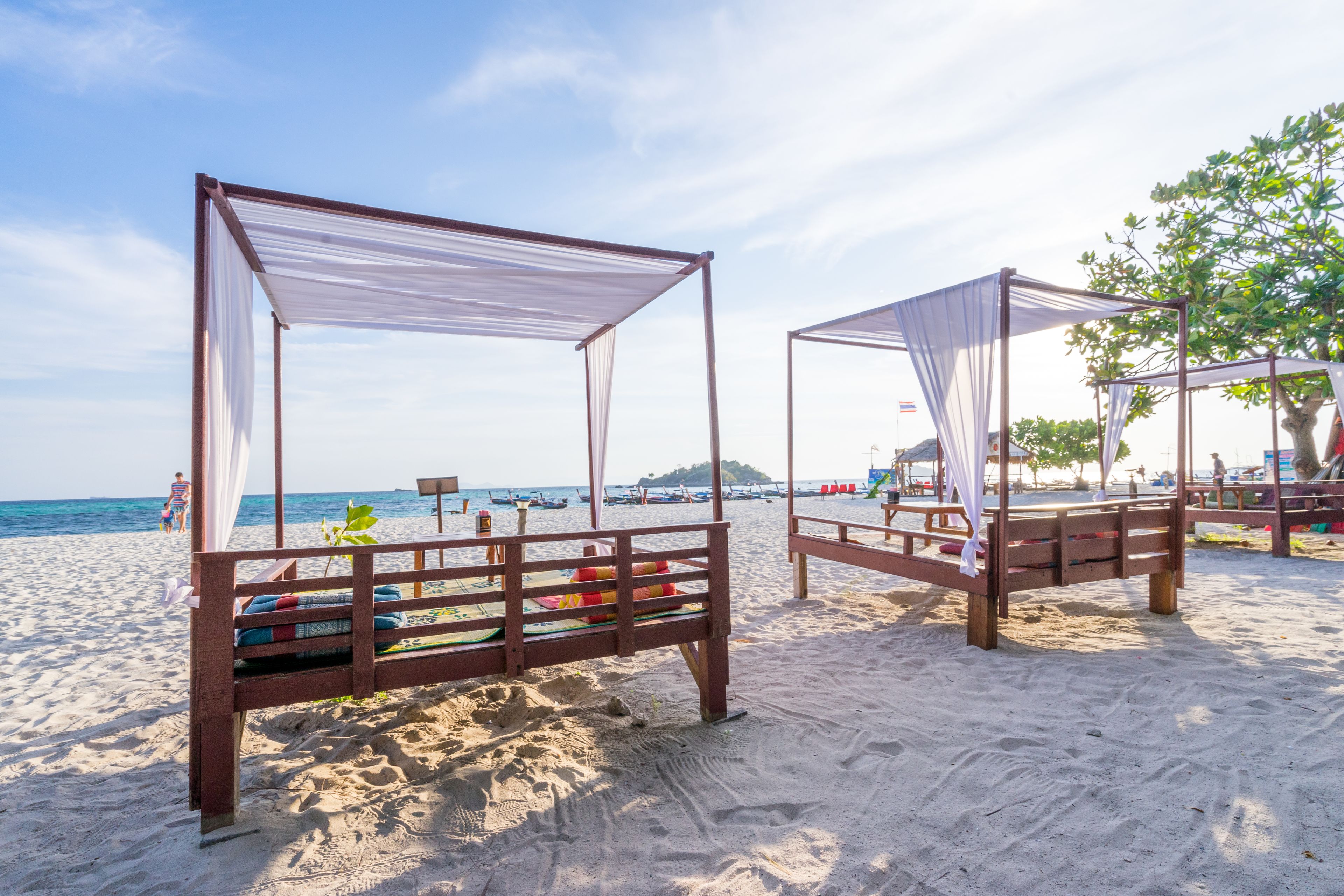 Two wooden beach beds with white canopies, set on a sandy beach with a view of the sea and distant buildings.