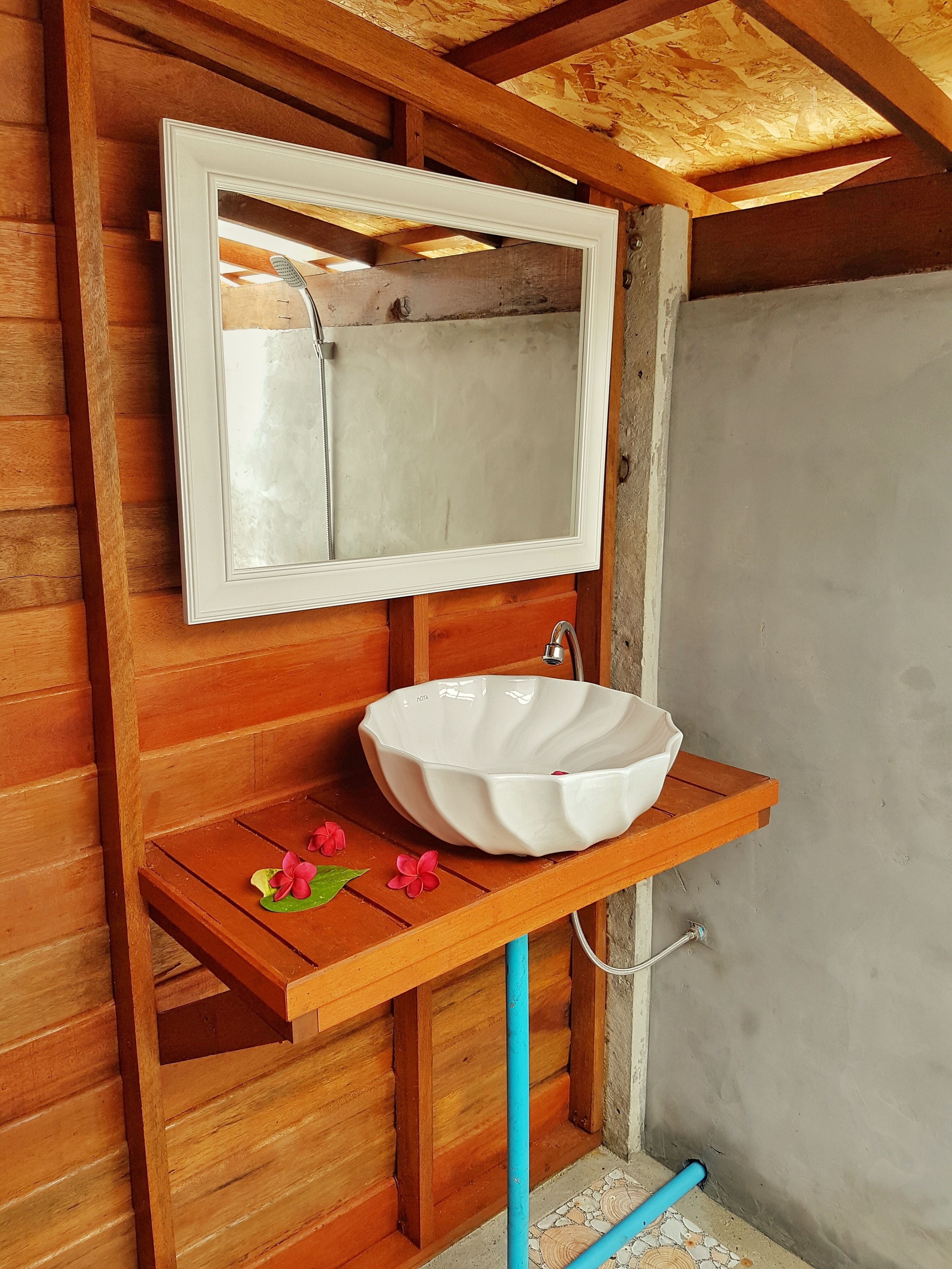 Interior de una cabaña de madera con un lavabo blanco y un espejo en un estante de madera, una tubería azul y una pared de concreto.