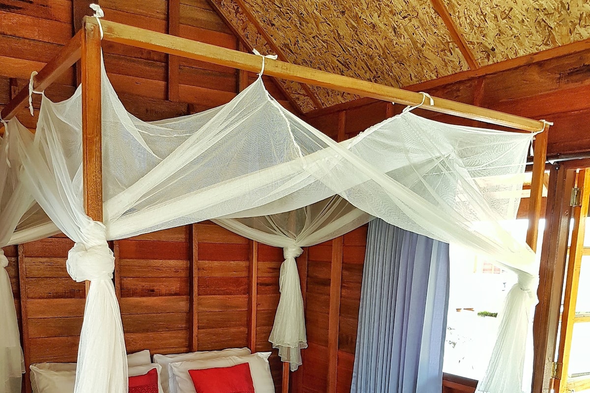 Quarto de cabana de madeira com cama de dossel, roupa de cama branca e toalha azul no chão.