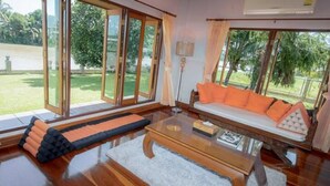 A spacious living room with a large glass door leading to a garden, a sofa with orange cushions, and a wooden coffee table.