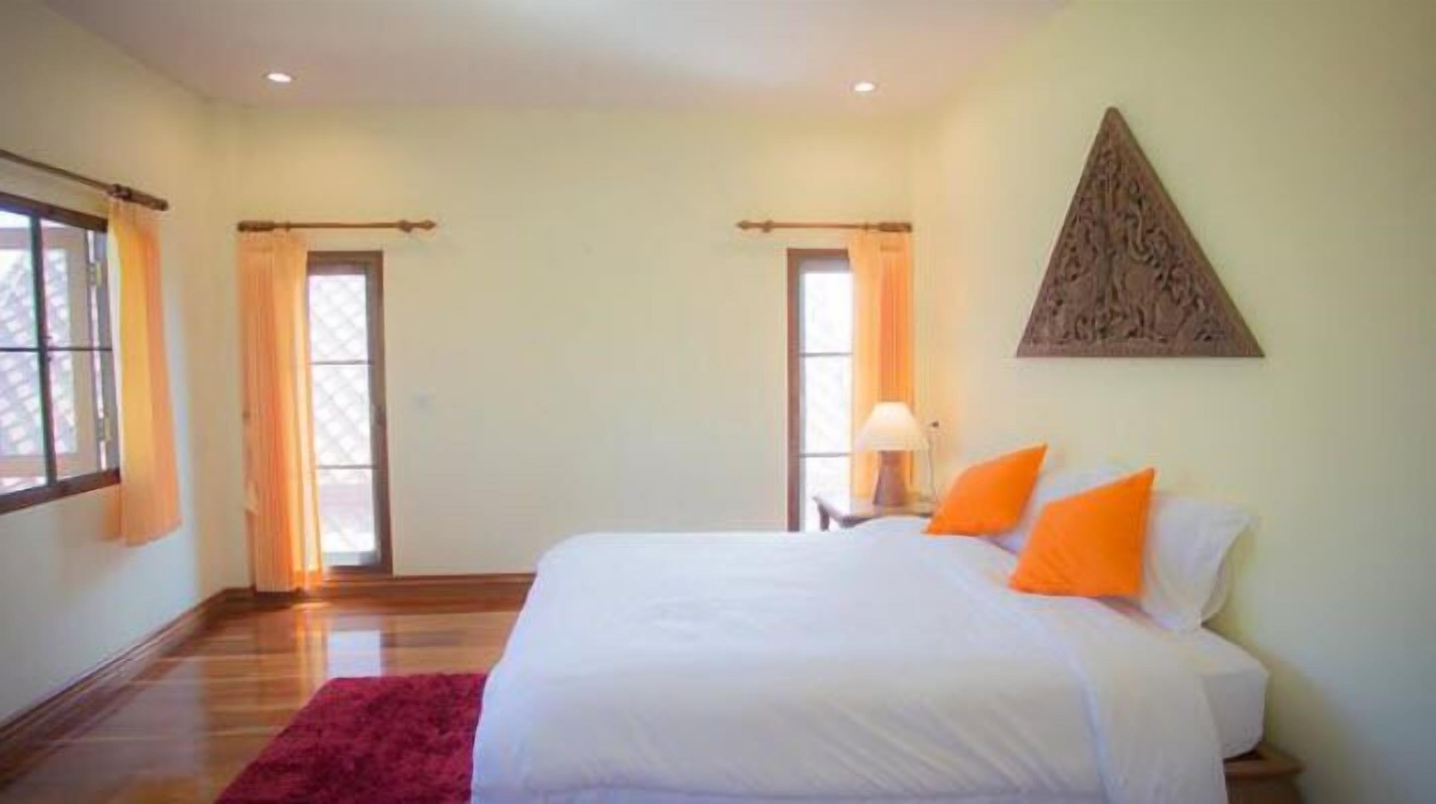 A bedroom with a large bed, two windows with curtains, a wooden floor, and a wall-mounted artwork.