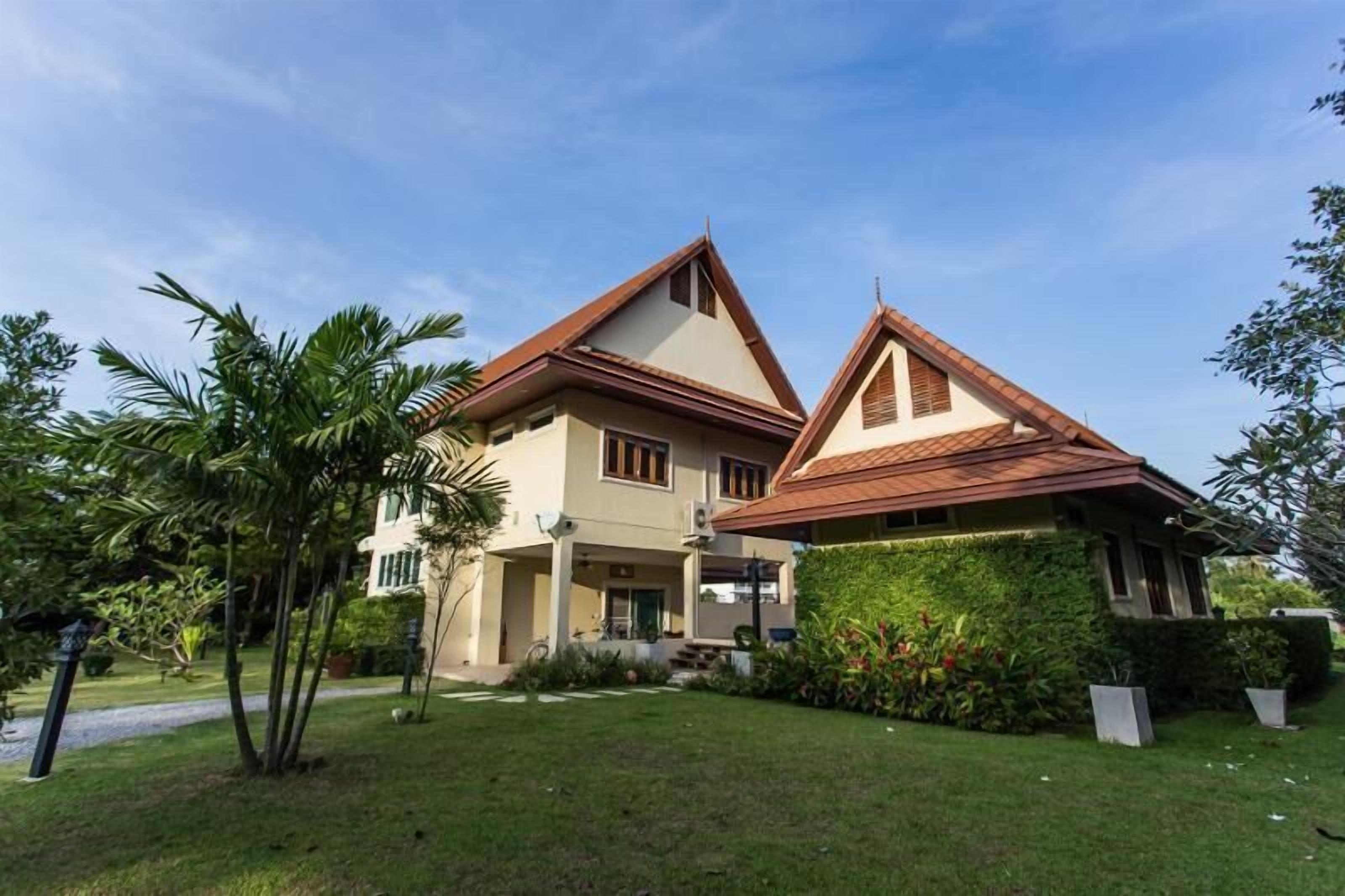 Uma casa de dois andares com telhado inclinado, varanda coberta e um jardim bem cuidado com palmeiras e arbustos.