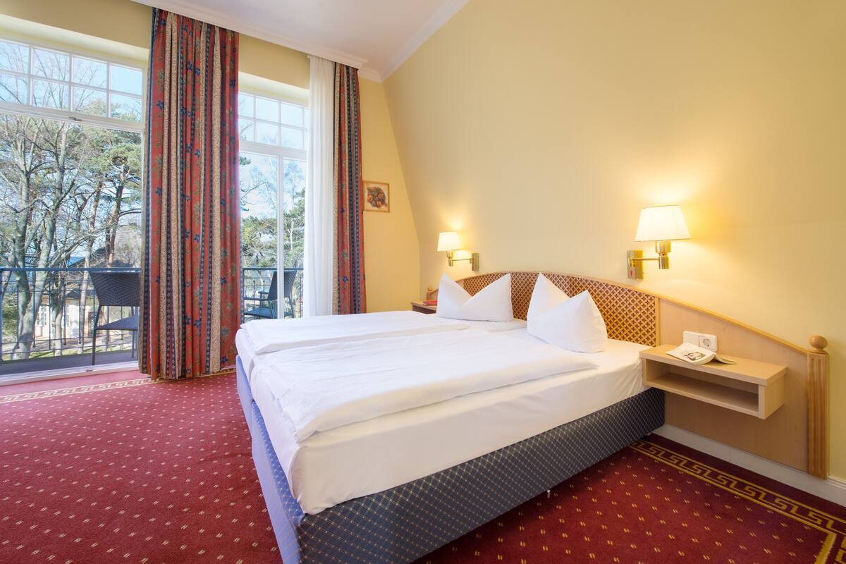 Ein Hotelzimmer mit einem großen Bett, zwei Nachttischlampen, einem kleinen Nachttisch und Blick auf Bäume durch das Fenster.