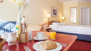 Een hotelkamer met een bed, een nachtkastje met een lamp, een ontbijttafel met een croissant, een kop koffie en een glas sinaasappelsap.