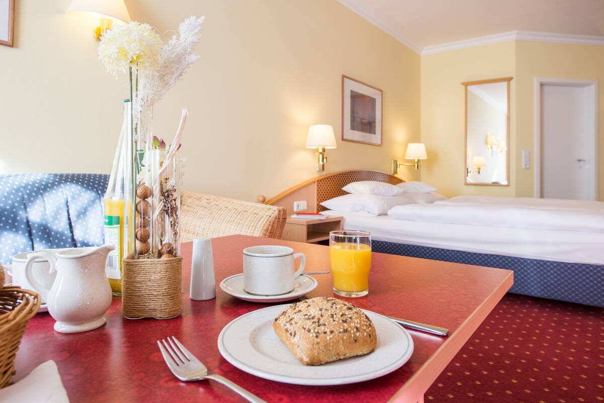 Ein Hotelzimmer mit Bett, Nachttisch mit Lampe, Frühstückstisch mit Croissant, einer Kaffeetasse und einem Glas Orangensaft.