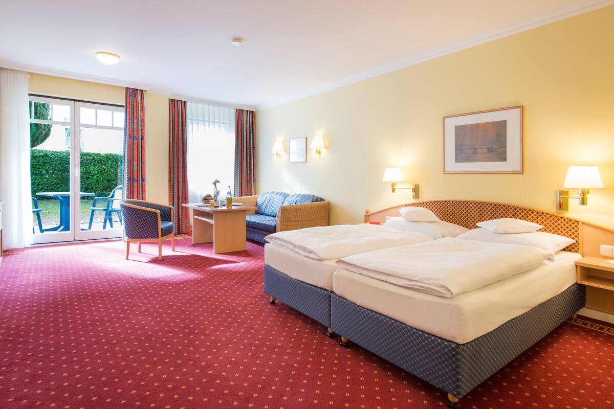 Ein Hotelzimmer mit Doppelbett, Schreibtisch mit Stuhl, einer Sitzecke und Blick ins Freie durch eine Schiebetür.