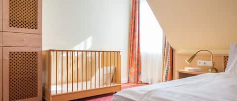 A room with a crib, a bed, a nightstand, and a lamp.