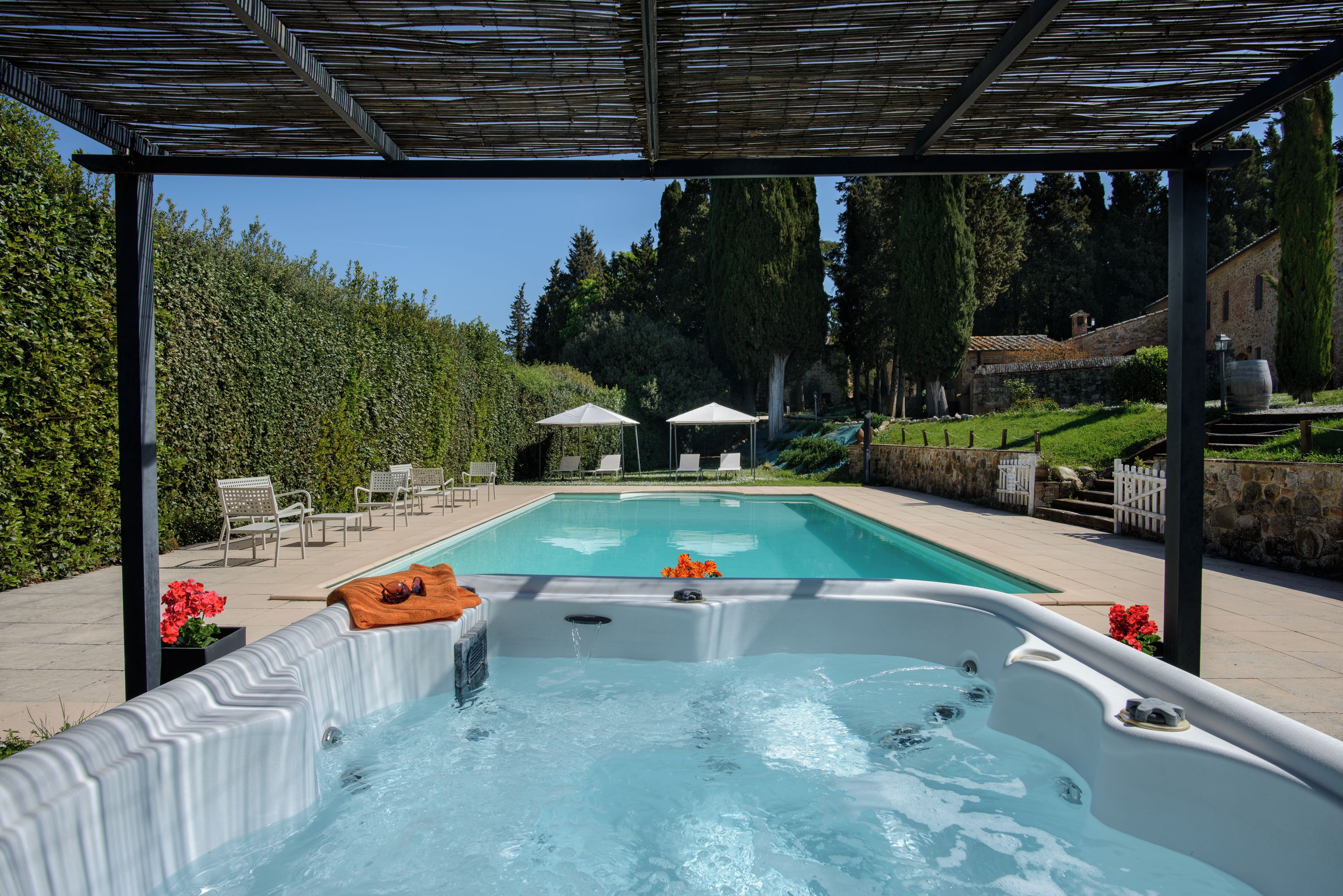 A swimming pool with a hot tub, surrounded by a pergola, chairs, and a hedge.