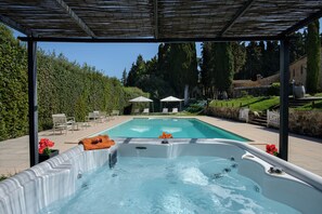 Una piscina con idromassaggio, circondata da un gazebo, sedie e un siepe.