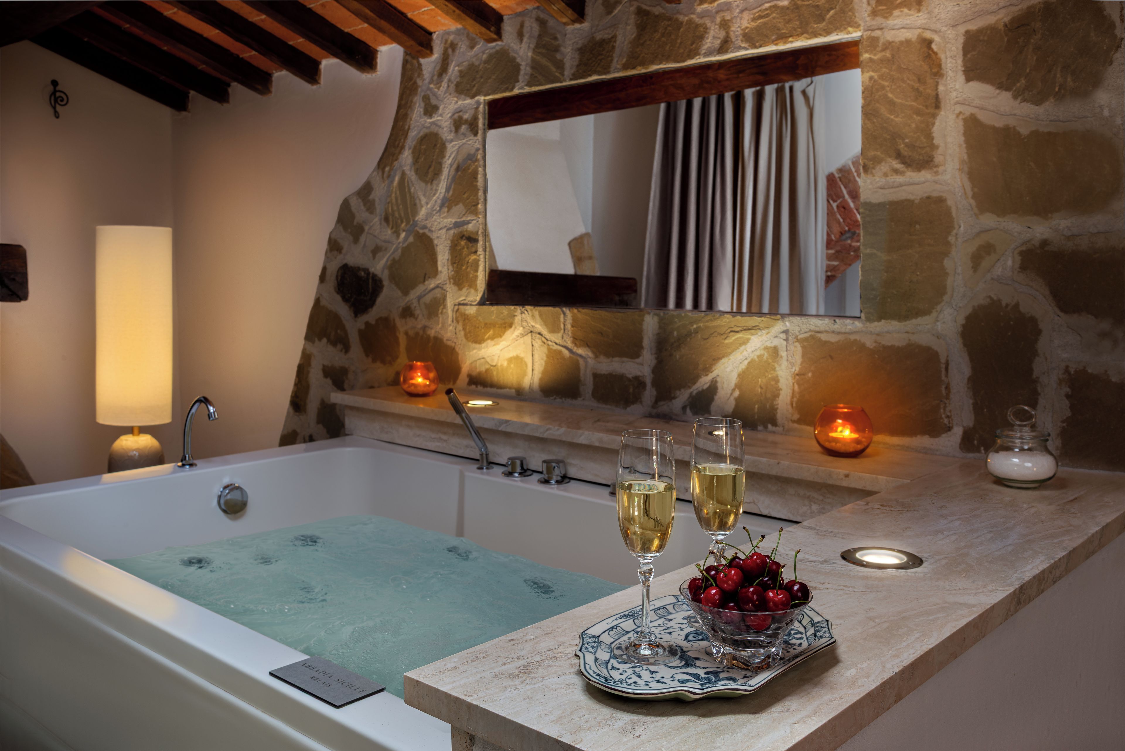 A bathroom with a bathtub, a mirror, and a countertop with two glasses of white wine and a bowl of cherries.