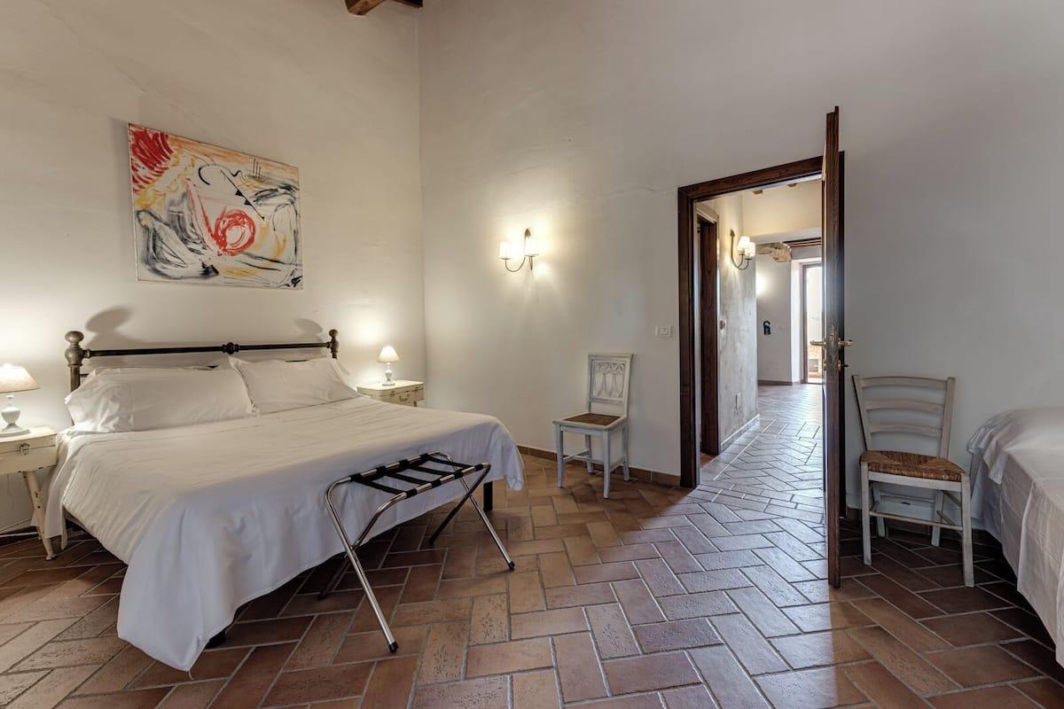 A bedroom with a bed, bedside tables, a chair, a painting, and a hallway leading to another room.