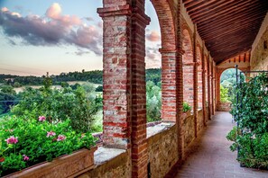 A covered corridor with brick columns, potted plants, and a view of a lush landscape. - Abbadia Sicille Relais (Trequanda)