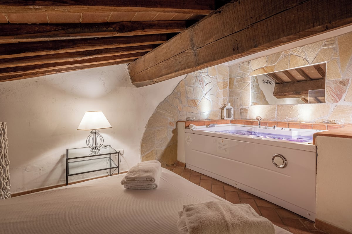 A bedroom with a stone wall, a wooden ceiling, a bed with white linens, a bedside table with a lamp, and a large bathtub.