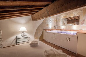 A bedroom with a stone wall, a wooden ceiling, a bed with white linens, a bedside table with a lamp, and a large bathtub.
