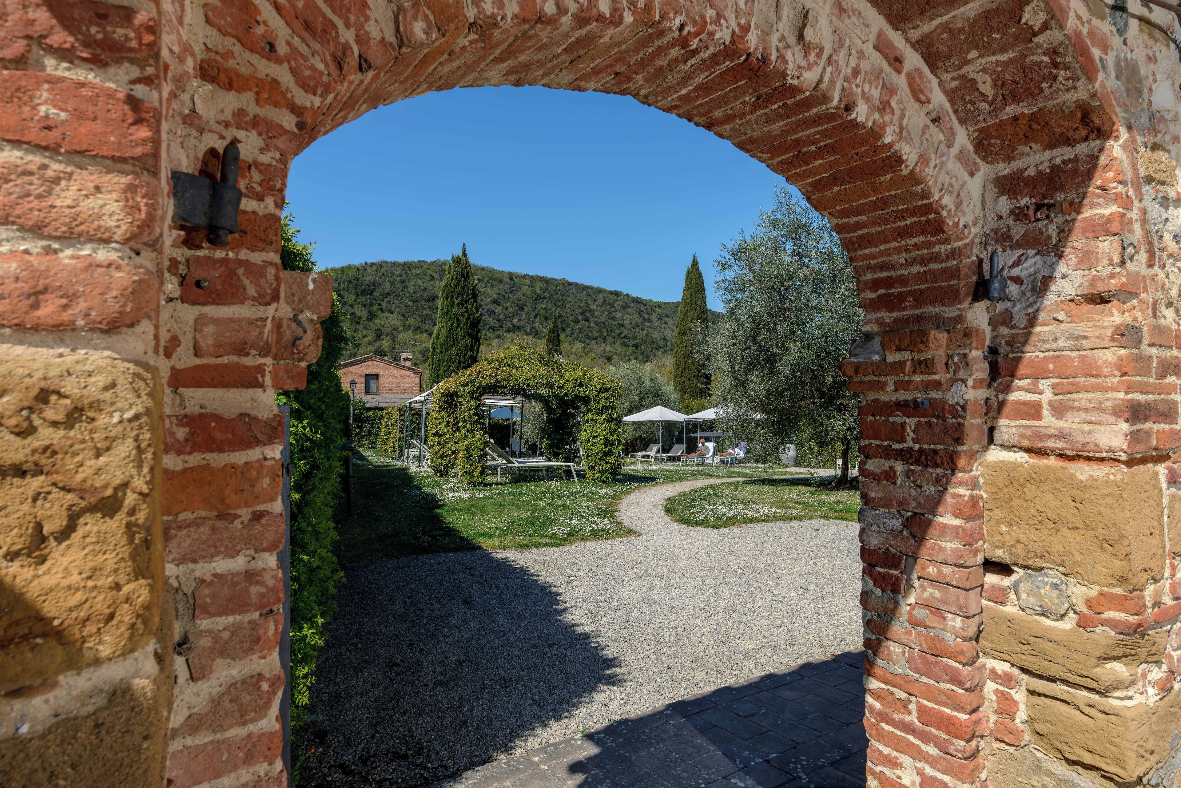 Une entrée en briques en forme d’arche mène à une cour intérieure avec un chemin de gravier, un bâtiment doté d’un toit en pente et une colline en arrière-plan.