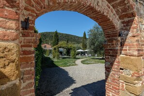 An arched brick entrance leading to a courtyard with a gravel path, a building with a sloped roof, and a hill in the background. - Abbadia Sicille Relais (Trequanda)