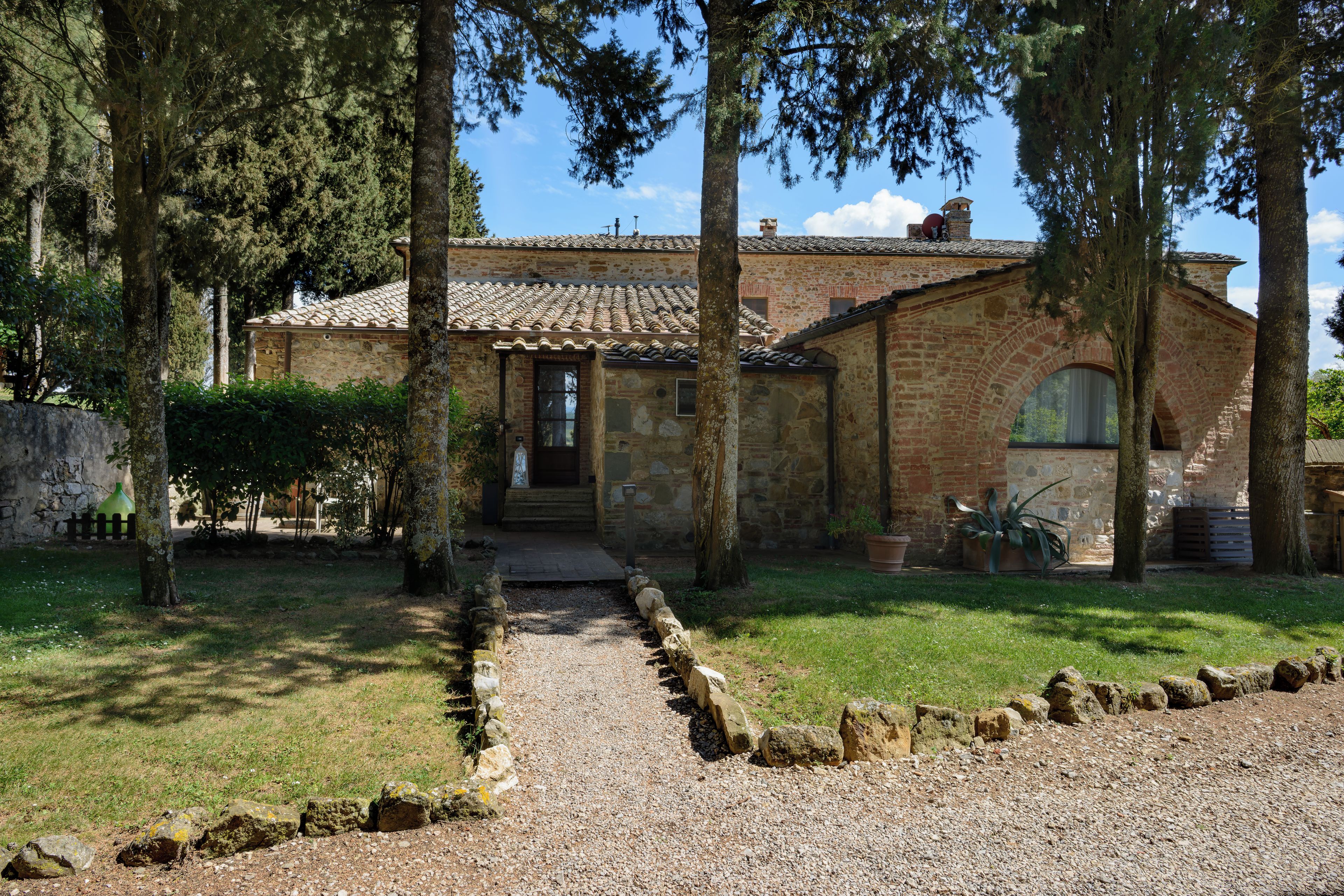 A rustic stone building with a tiled roof, surrounded by trees and a gravel path leading to the entrance.