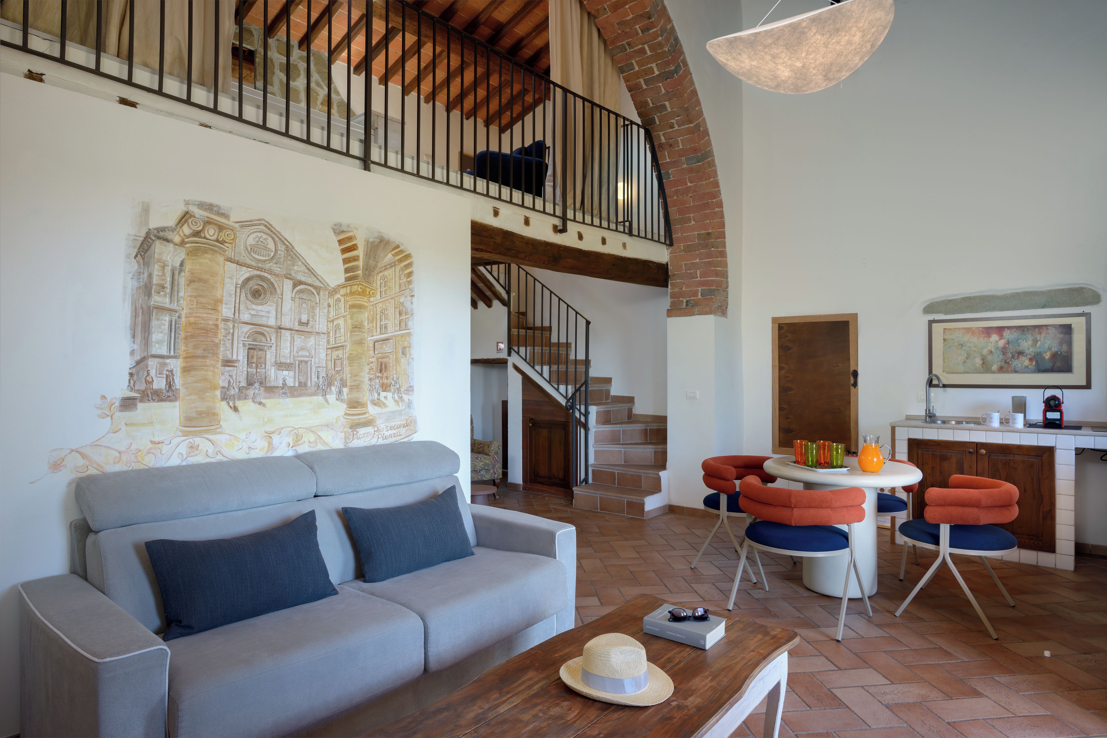 A living room with a sofa, a coffee table, a mural, a staircase, and a kitchen area.
