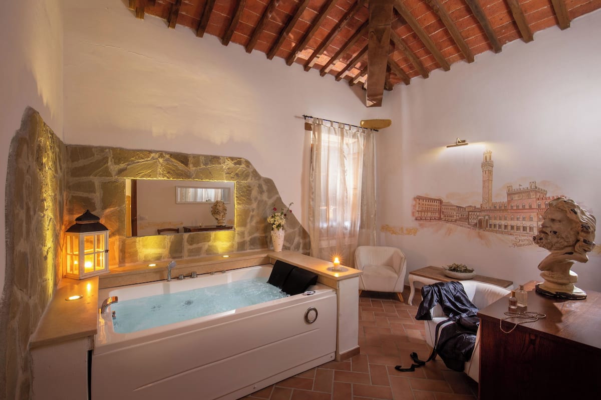 A bathroom with a stone wall, a bathtub, a mirror, a chair, and a decorative bust.