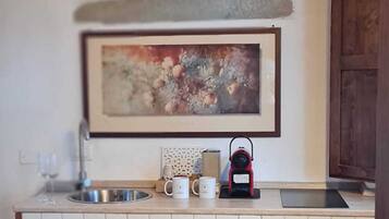 Uma cozinha com painel de azulejos brancos, um armário de madeira, uma pia de aço inoxidável, uma bancada com chaleira, cafeteira e um quadro emoldurado sobre a pia.