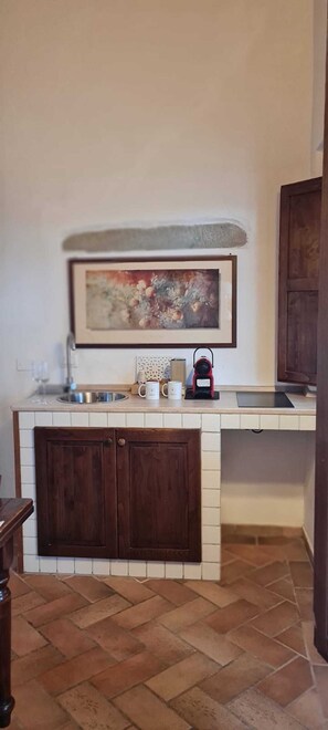 Una cucina con schienale in piastrelle bianche, un armadietto in legno, un lavello in acciaio inossidabile, un piano di lavoro con bollitore, macchina del caffè e un quadro incorniciato sopra il lavello.