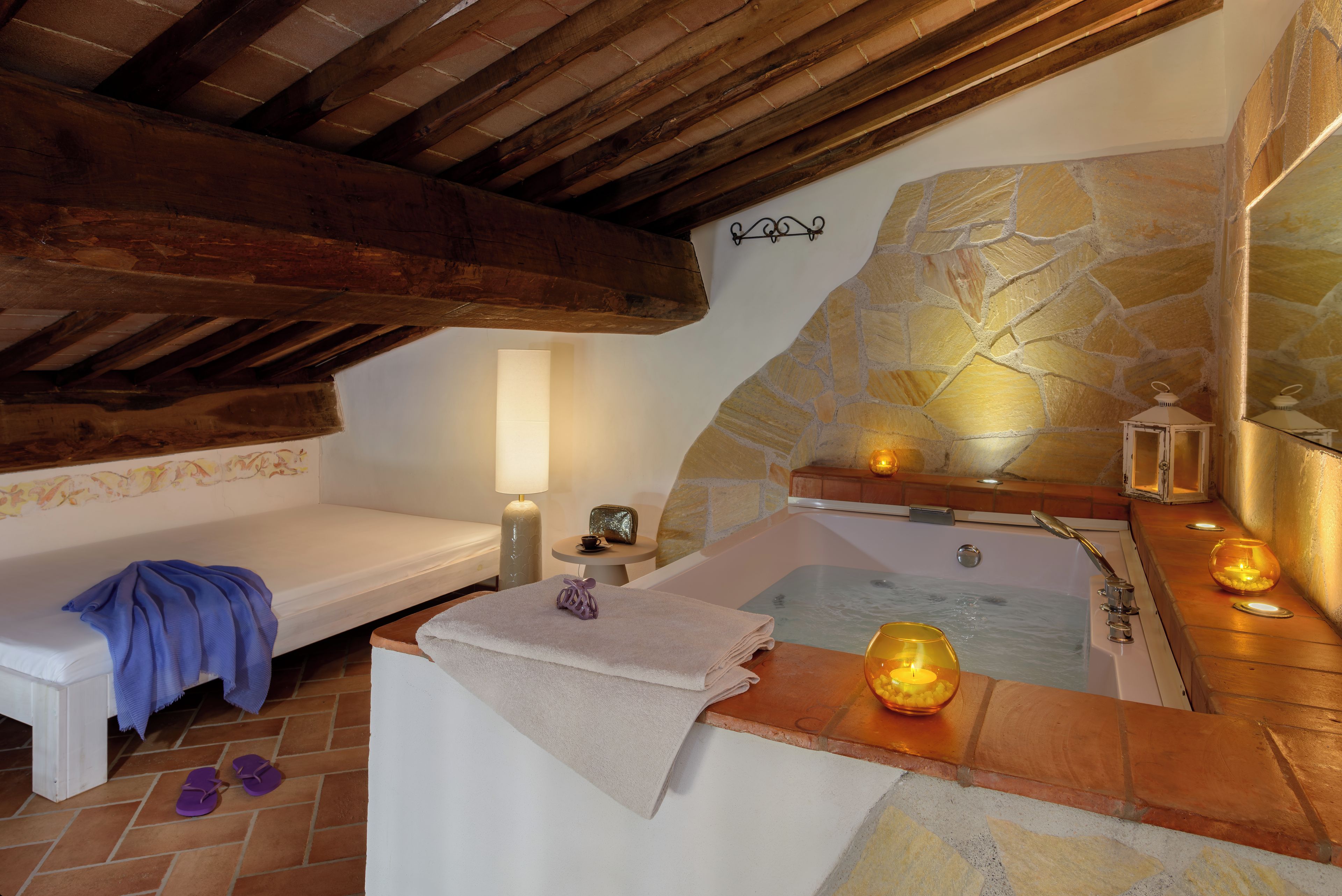 Un cuarto de spa con pared de piedra, un banco de madera, una bañera y una mesita con una lámpara y una taza.