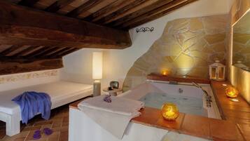 Un cuarto de spa con pared de piedra, un banco de madera, una bañera y una mesita con una lámpara y una taza.