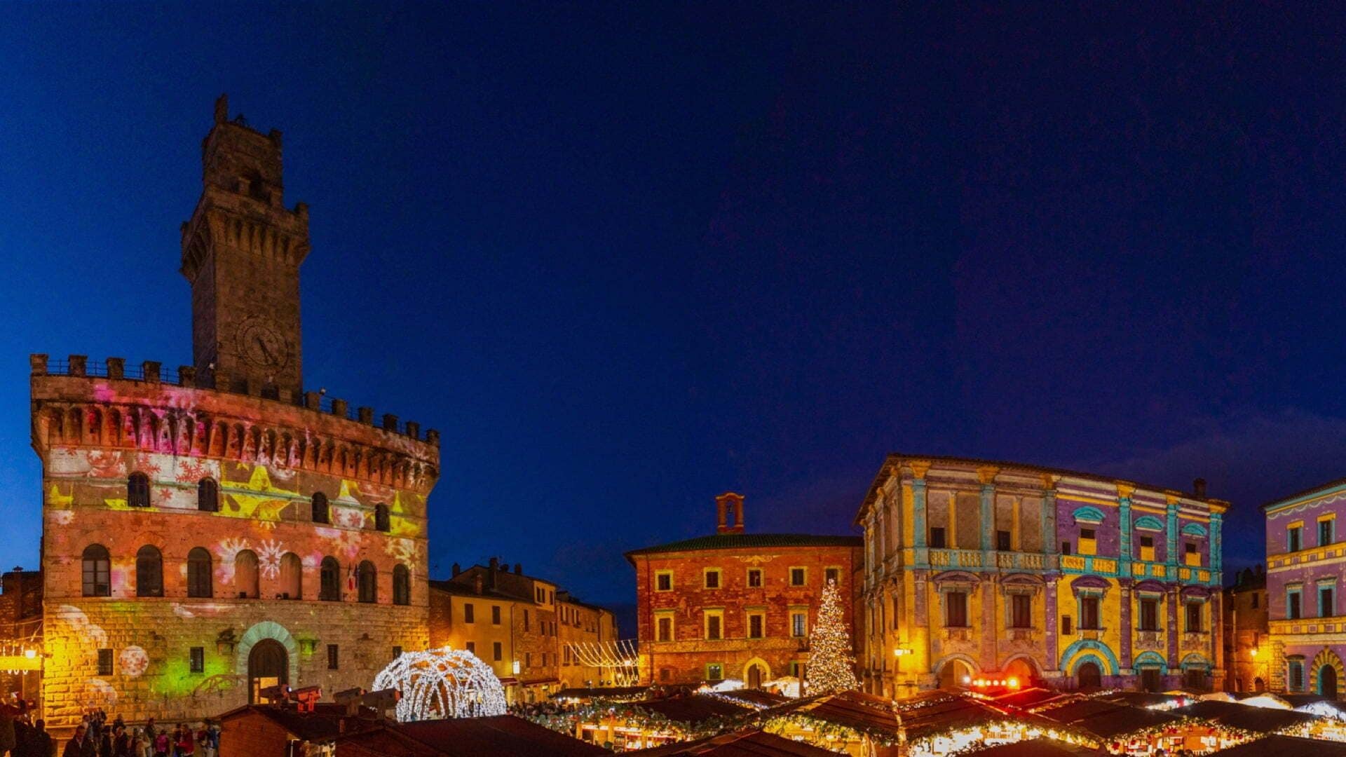 A historic square with a prominent tower and illuminated buildings, suggesting a festive atmosphere.