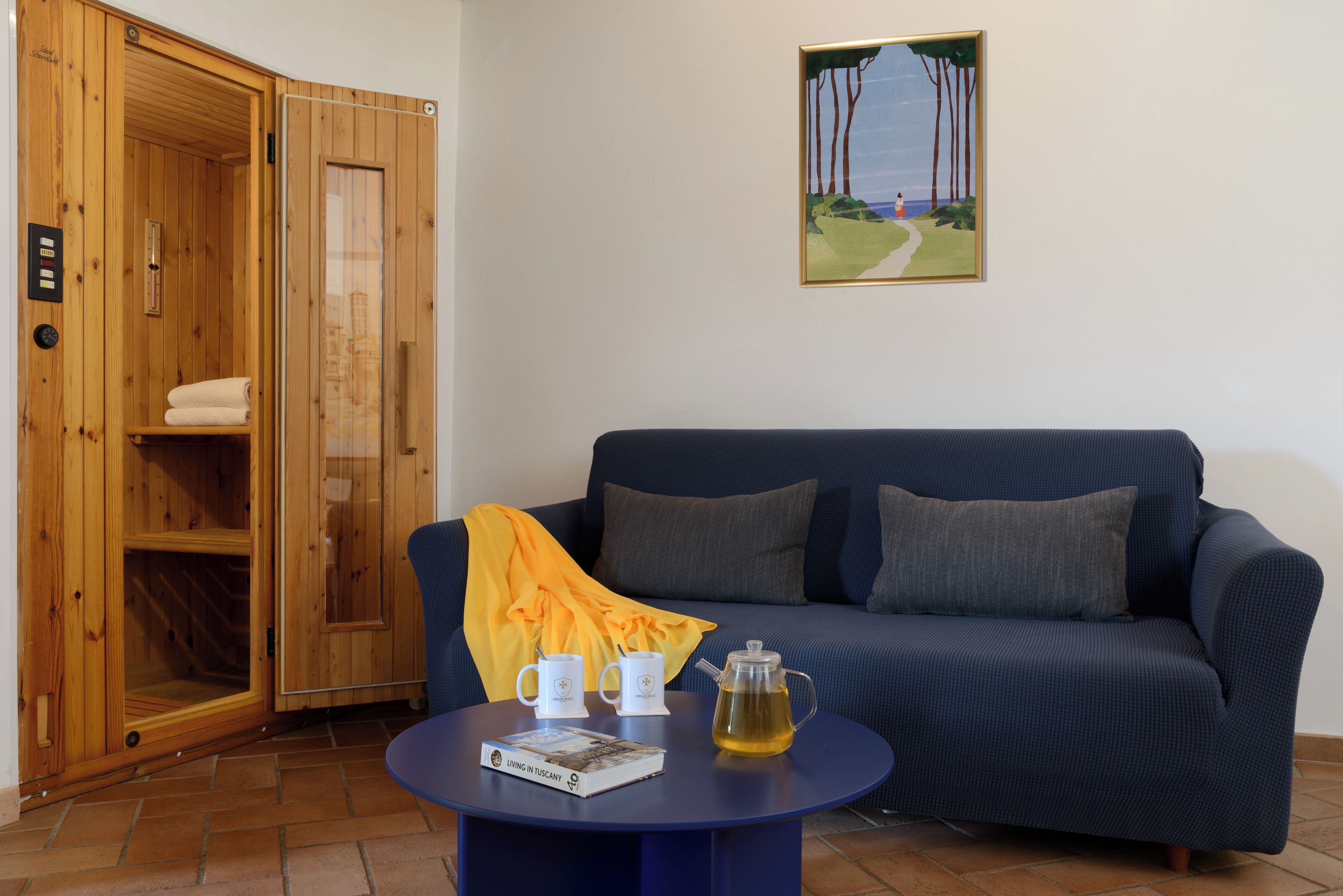A room with a wooden sauna, a blue sofa, a table with a book and a teapot, and a painting on the wall.