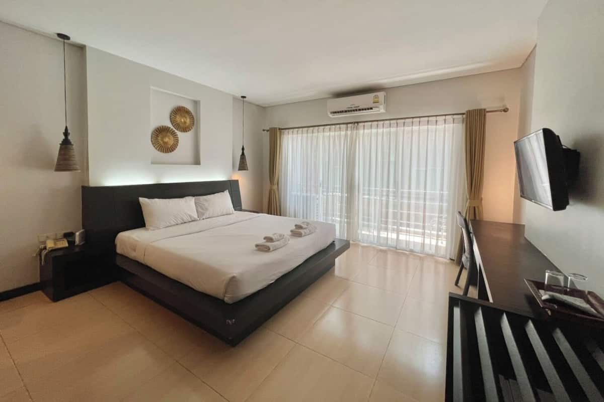 A modern hotel room with a large bed, a desk with a flat-screen TV, and a large window with sheer curtains.