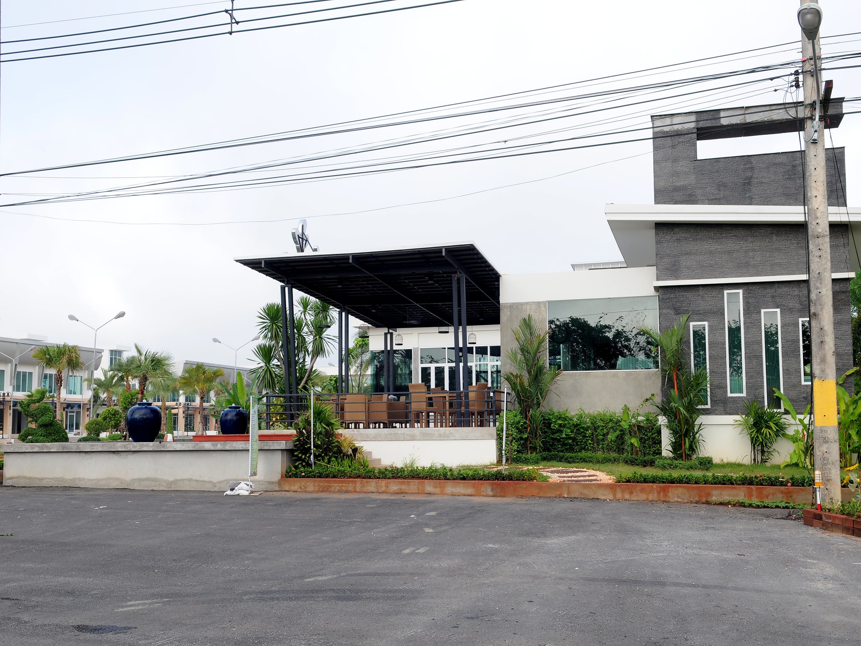 A modern building with a covered outdoor seating area, surrounded by greenery and palm trees.