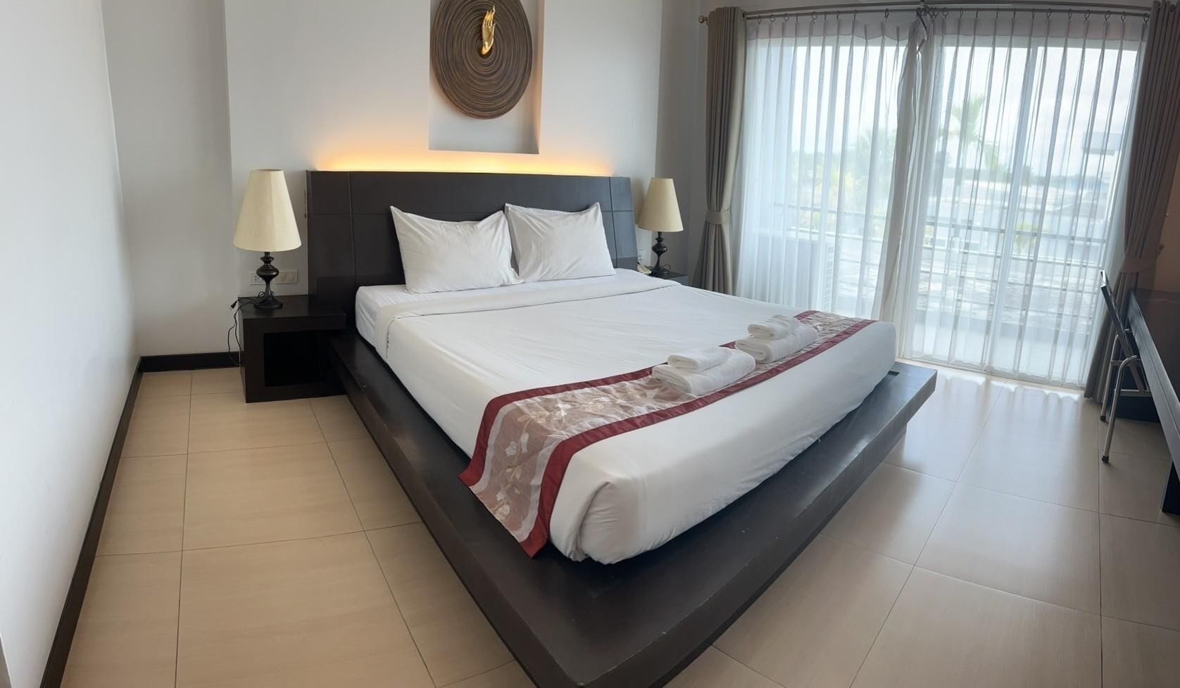 A neatly made bed with white linens and a red and white patterned throw, two bedside lamps, a dark headboard, and a large window with sheer curtains.