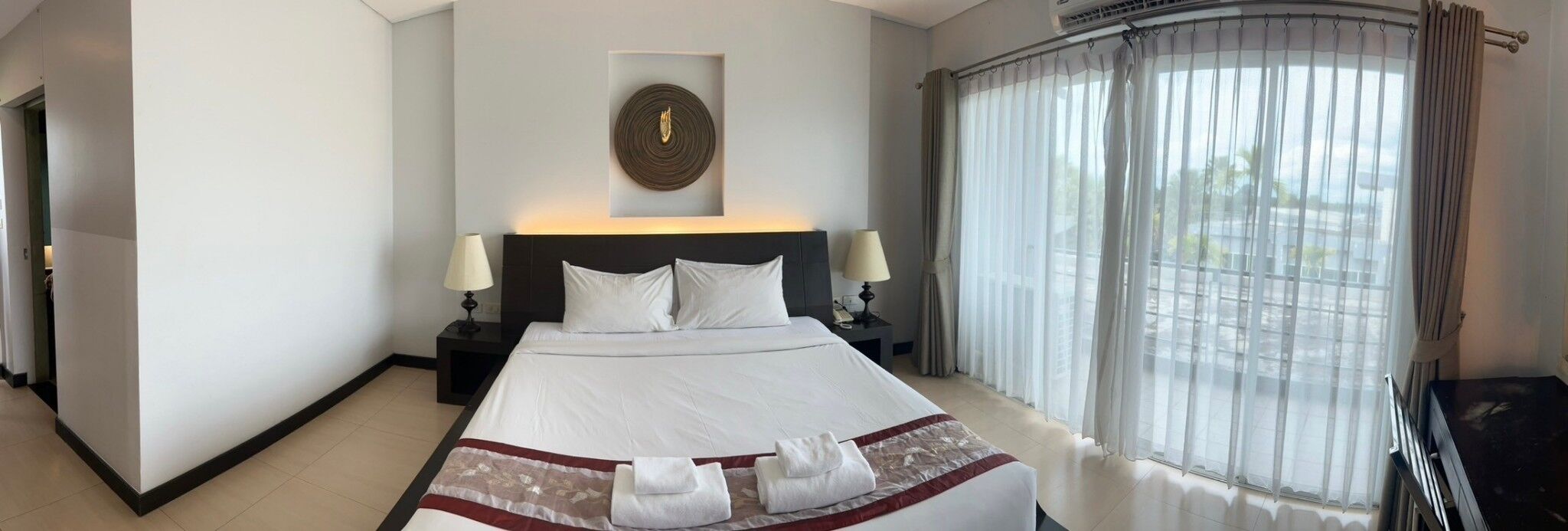Una habitación de hotel con una cama grande, dos mesitas de noche con lámparas, una obra de arte colgada en la pared y vistas a través de cortinas de gasa.