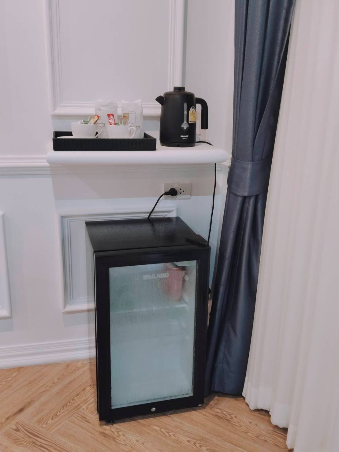 Un refrigerador compacto con puerta de vidrio, una cafetera en un estante y un enchufe en la pared.