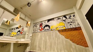 A bathroom with a large mirror, a sink, and a wall with cartoon characters. - The Hive Hatyai Hostel (Hat Yai)