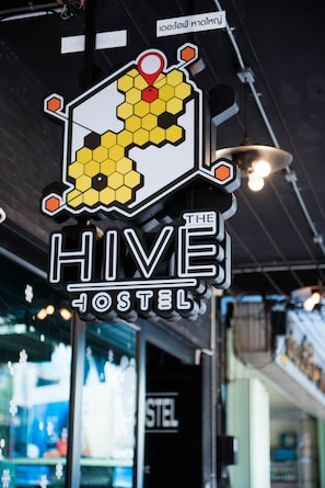 A sign for "The Hive Hostel" with a hexagonal logo featuring a honeycomb pattern and a red location pin. - The Hive Hatyai Hostel (Hat Yai)