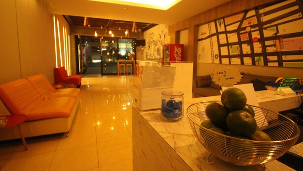 A hotel lobby with a sofa, a coffee machine, a Coca-Cola fridge, a bulletin board, and a bowl of fruit. - The Hive Hatyai Hostel (Hat Yai)