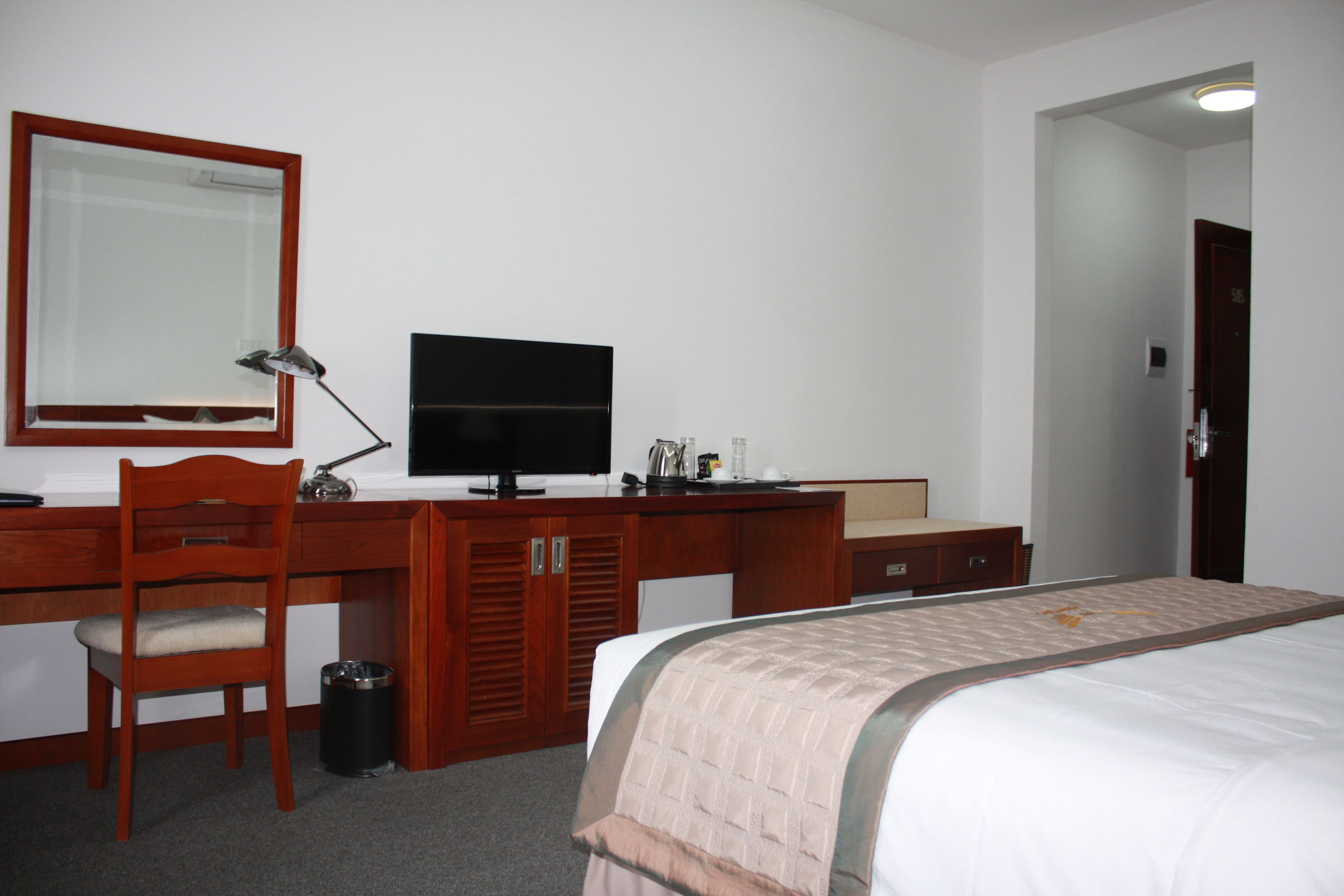 Quarto de hotel com escrivaninha de madeira, televisão, cama com manta bege e espelho na parede.