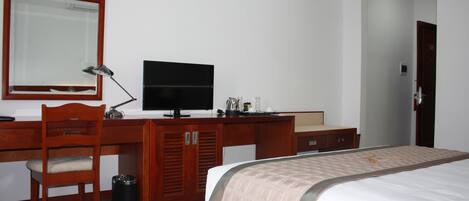 Quarto de hotel com escrivaninha de madeira, televisão, cama com manta bege e espelho na parede.