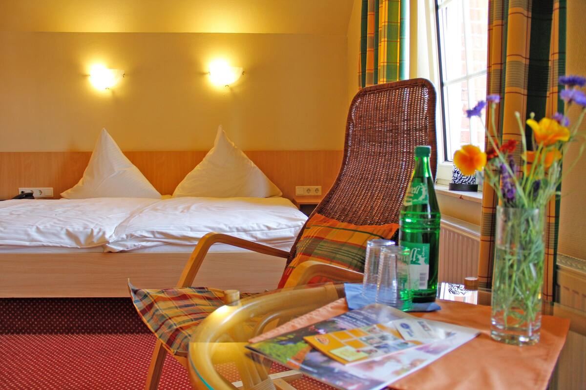 A hotel room with a bed, two pillows, a wicker chair, a table with a bottle, a glass, and a vase with flowers.