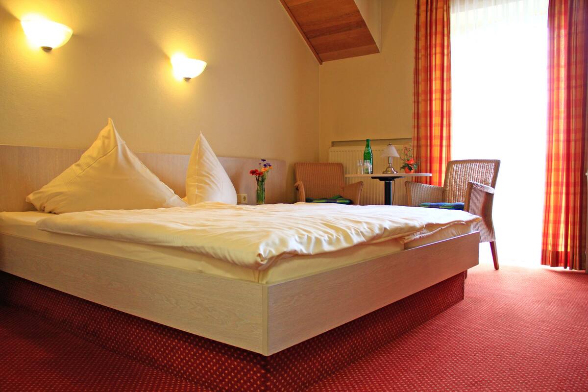 A large bed with white linens, two wall-mounted lights, a small table with a lamp, and a chair.
