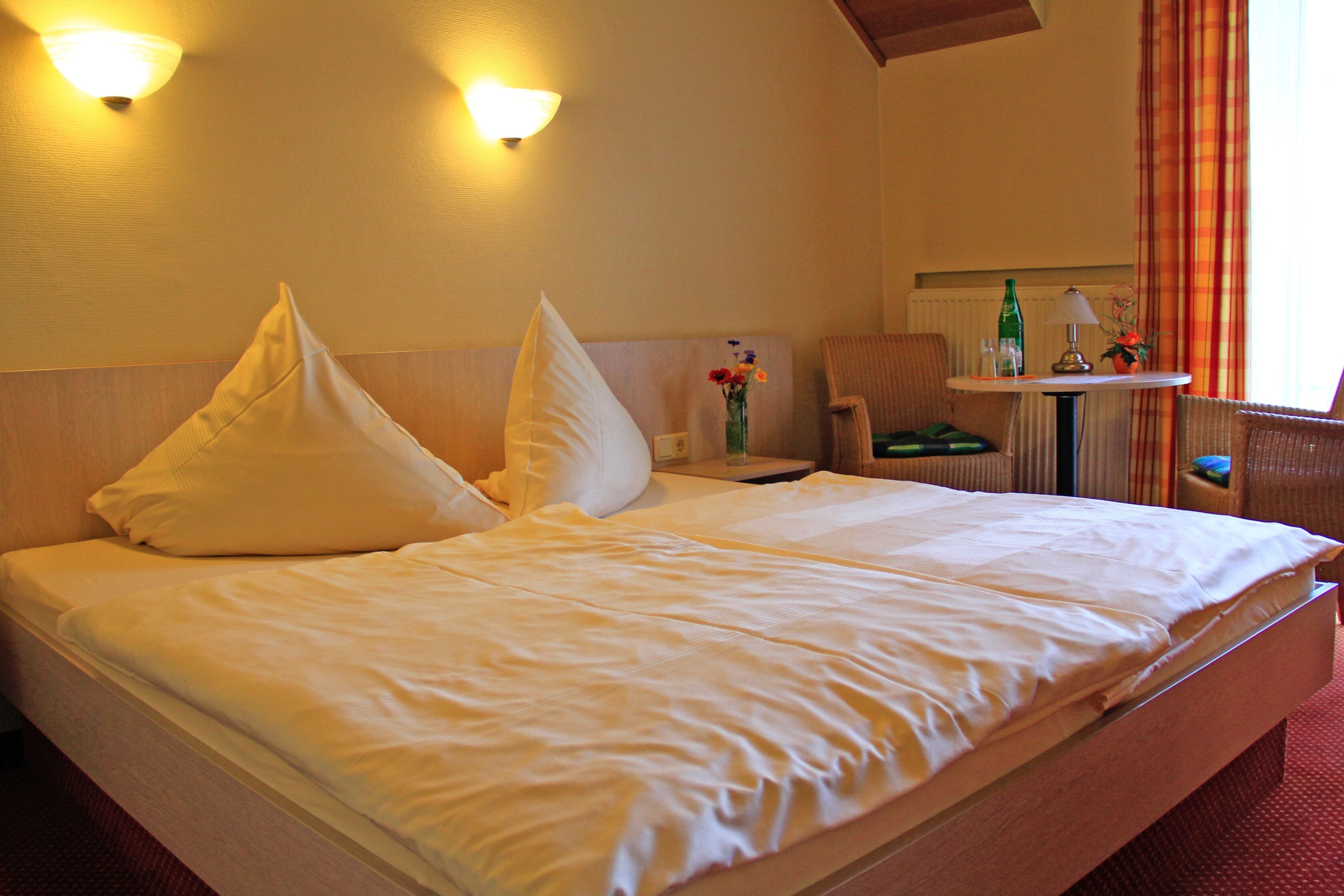 Una cama bien hecha con sábanas y almohadillas blancas, una mesita de noche con una lámpara y un florero, y una silla en una habitación de hotel.