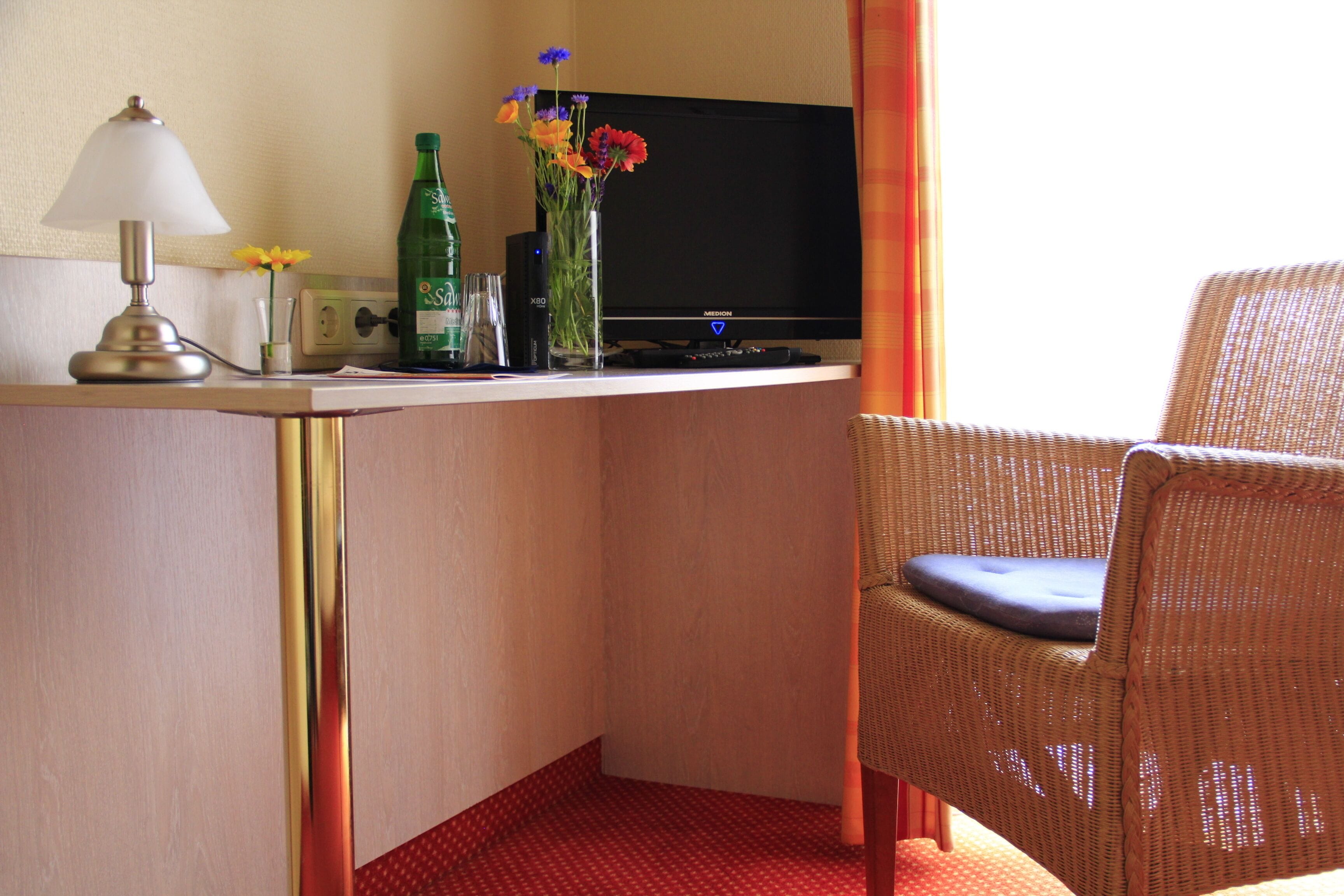 Habitación de hotel con escritorio, televisor, lámpara, una botella, un florero con flores y una silla de mimbre.