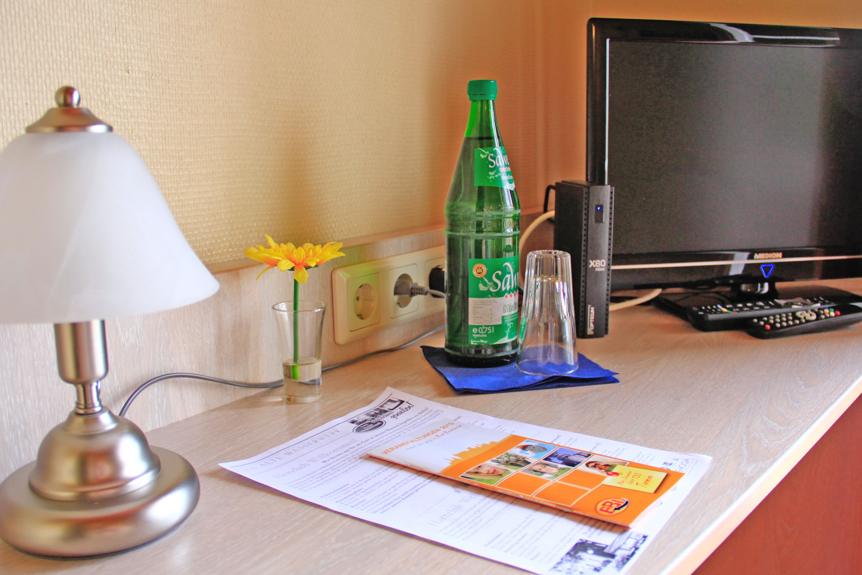 A hotel desk with a lamp, a bottle of soda, a glass, a remote control, and a brochure.