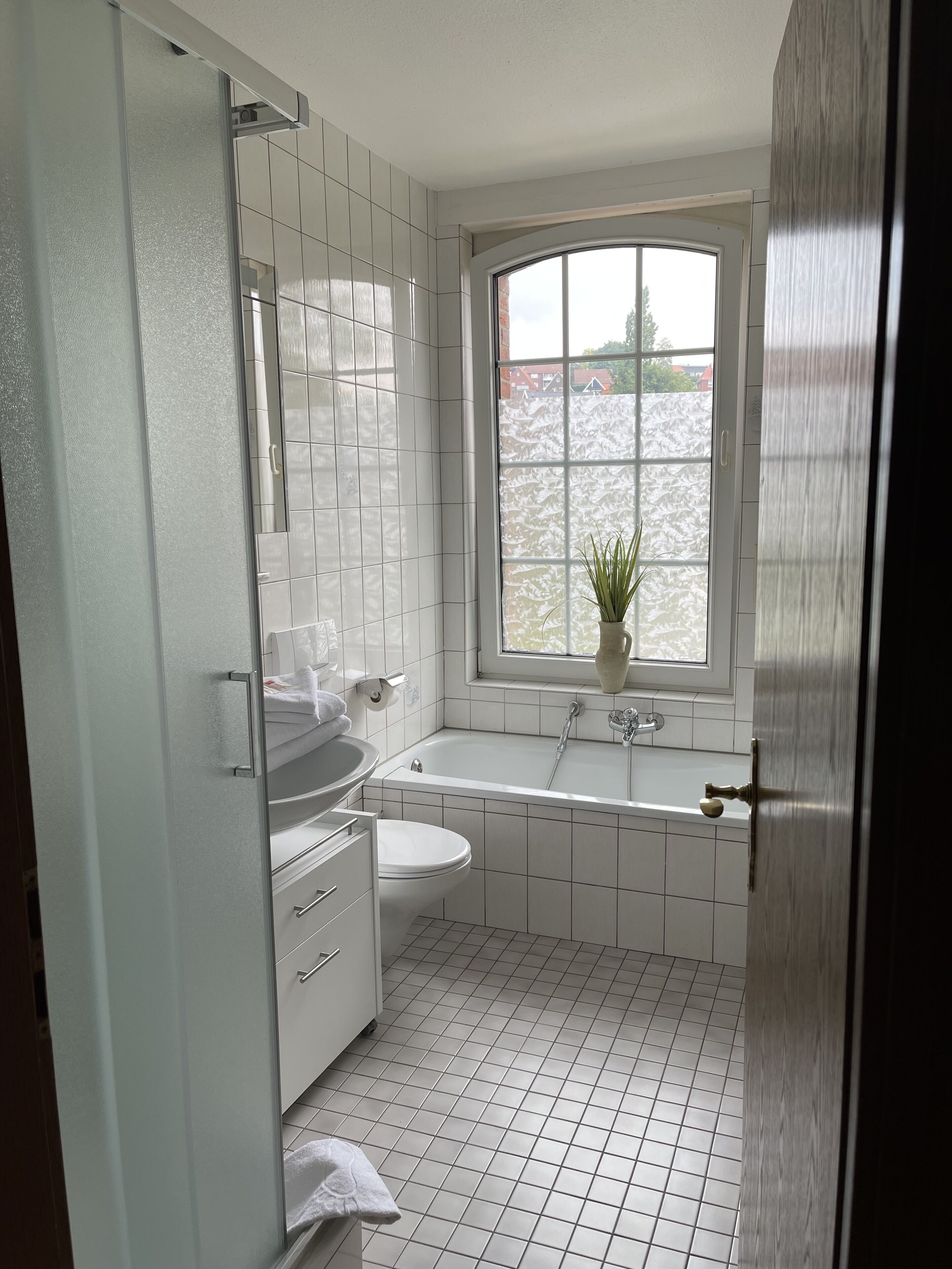 Un baño con lavamanos, inodoro y bañera blancos, una ventana con vistas a árboles y una planta en maceta en el alféizar.