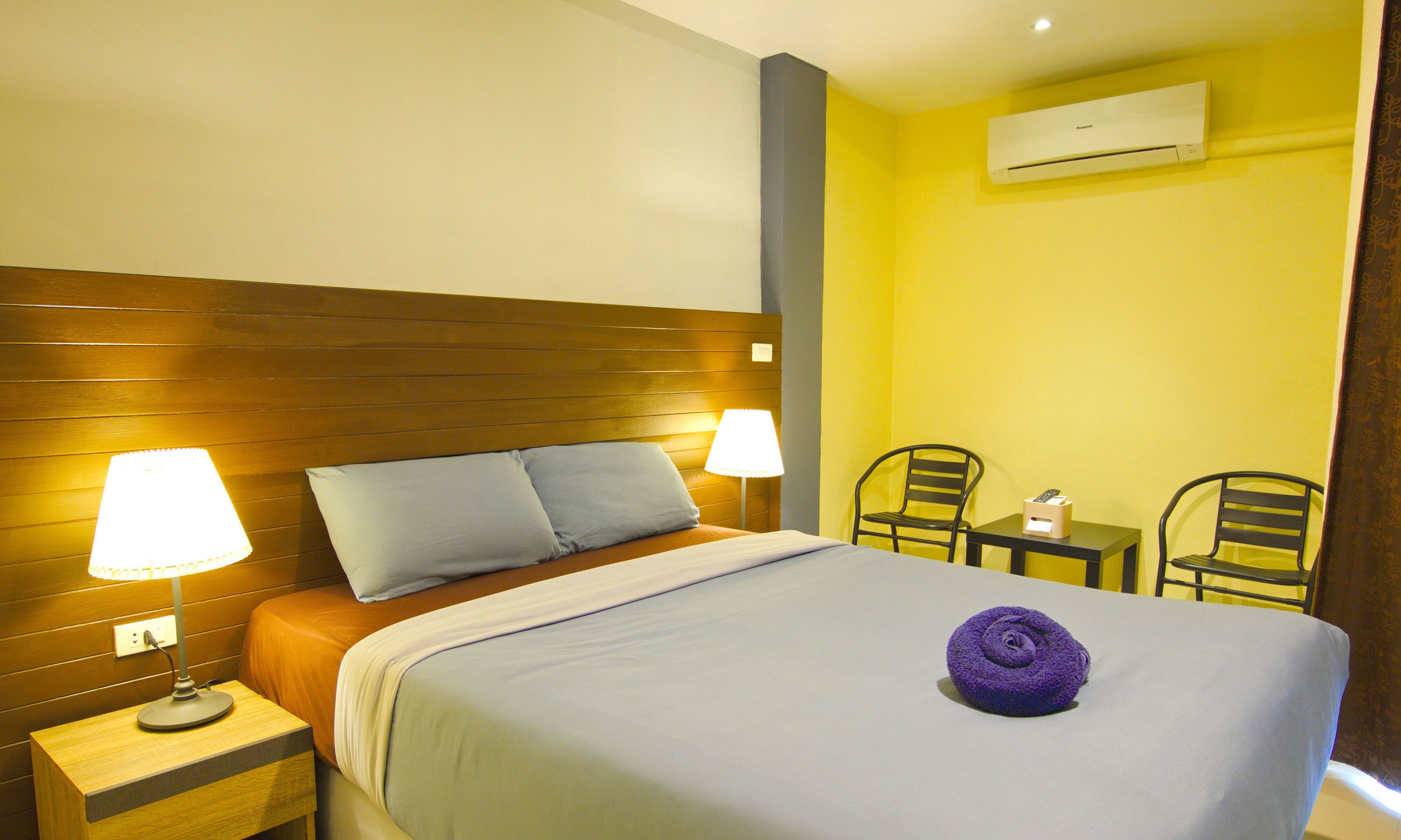 A hotel room with a wooden headboard, two bedside lamps, a bed with a blue bedspread, a purple towel, a small table with a remote control, two chairs, and an air conditioning unit on the wall.