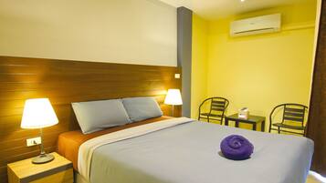 A hotel room with a wooden headboard, two bedside lamps, a bed with a blue bedspread, a purple towel, a small table with a remote control, two chairs, and an air conditioning unit on the wall.