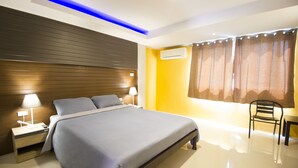 A modern hotel room with a large bed, bedside lamps, a wooden headboard, a window with curtains, and a small table with a chair. - The Avail (Ayutthaya)
