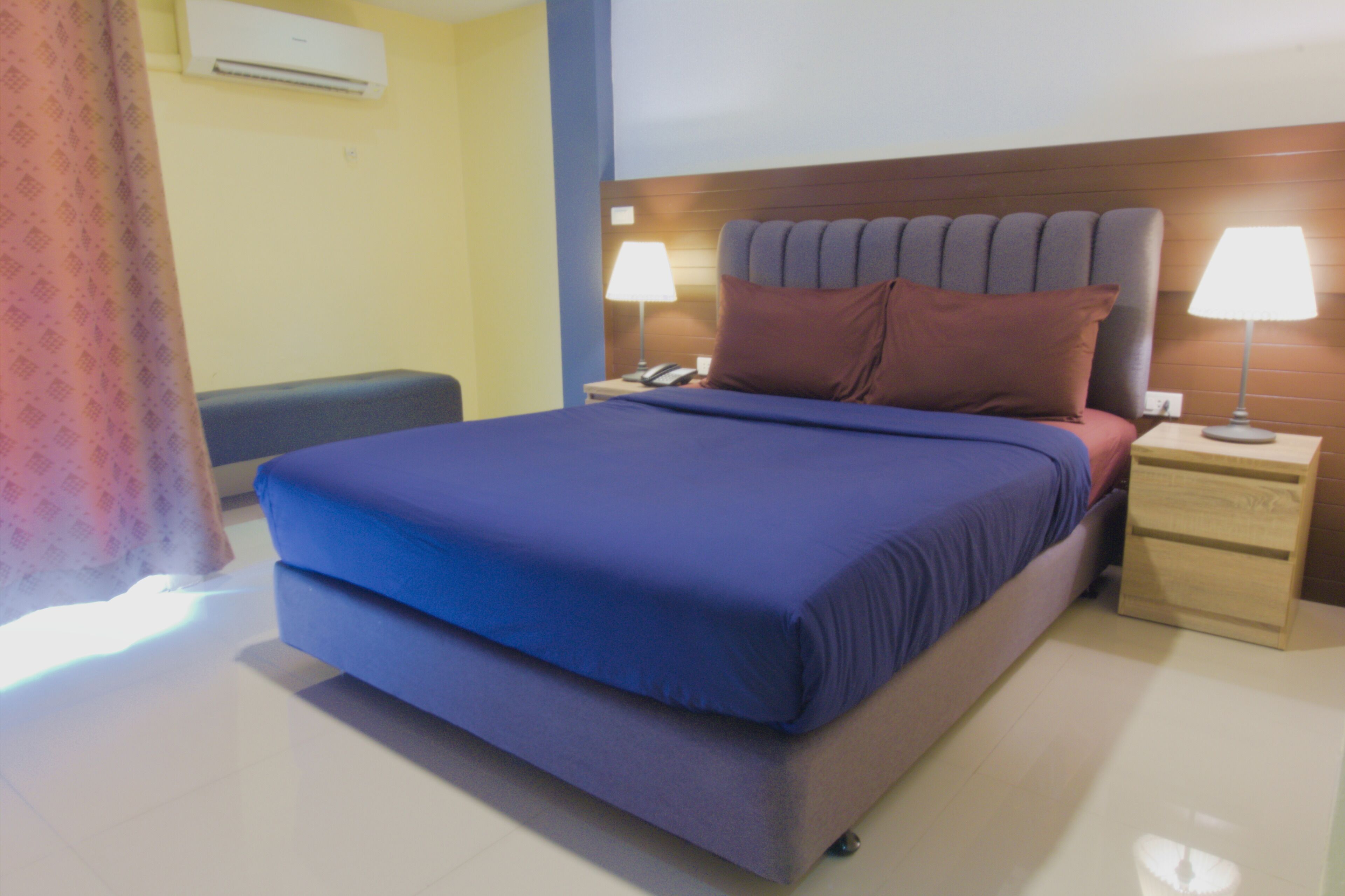 A hotel room with a bed, two bedside tables with lamps, a nightstand, and a wall-mounted air conditioner.